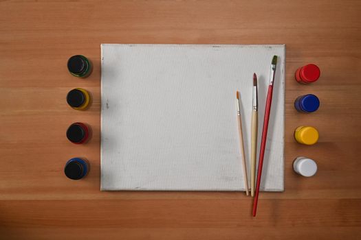 Flat lay blank canvas, watercolors and paint brushes on wooden background. Artist or designer workplace.