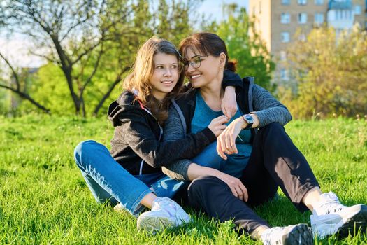 Happy mother and daughter hugging sitting on grass. Mom and girl resting together on lawn. Family, leisure, lifestyle, relationship, love, mother's day, motherhood concept