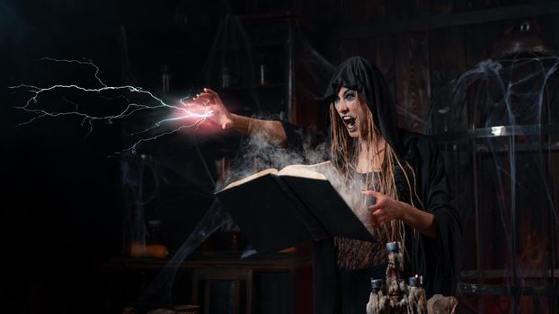 Halloween concept. Witch dressed black hood standing dark dungeon room use magic book for conjuring magic spell with lightning. Female necromancer wizard gothic interior put her hand forward screaming
