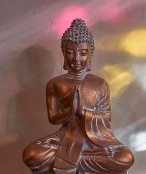 Golden buddha statuette with hands folded like the Indian Namaste greeting (Anjali mudra). He sits in pink and yellow light reflections coming from objects in the window.