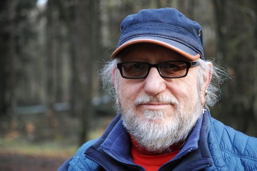 Portrait of a 61-year-old senior man wearing blue clothes and a cap. He has a beard and glasses with tinted lenses