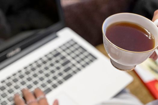 a girl with a cup of tea in her hand is typing on a laptop.Remote work on a computer. Distance education. Home office and freelance