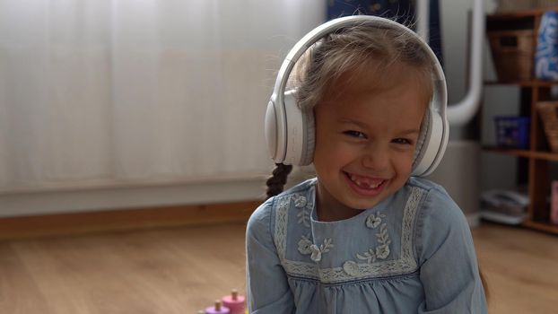 Child Listening Music In Big White Headphones. Happy Little Preschool Toothless Girl 5 Years Old Looking At Camera In Leaving Room Inside. Kid Smiles Laughs Shows Tongue At Home. Childhood, Education.