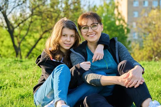 Happy mother and daughter hugging sitting on grass, looking at camera. Mom and girl resting together on lawn. Family, happiness, leisure, lifestyle, relationship love, mother's day, motherhood concept