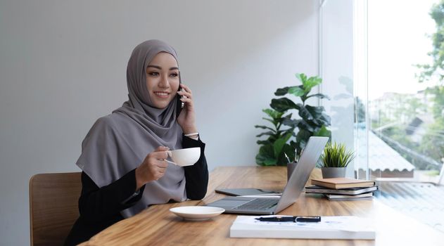 happy beautiful muslim female worker holding hot coffee mug sitting in office and using mobile cell phone viewing online news relaxing..