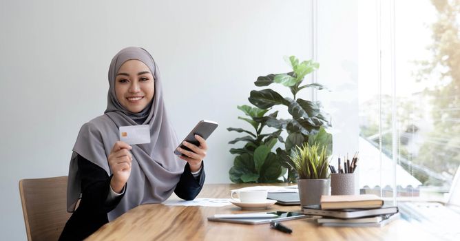 Beautiful young muslim woman with hijab sits at her office desk, holding a smartphone and credit card. Online payment, internet banking, online shopping concept. Close-up image.