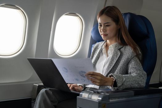 Asian young woman using laptop with financial report sitting near windows at first class on airplane during flight, Traveling and Business concept.