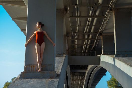 Beautiful young strong healthy woman ballerina gymnast in red leotard posing as model on bridge girder and blue sky and looking into the distance.