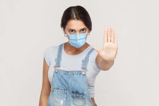 Stop. Portrait of angry young brunette woman with surgical medical mask in denim overalls standing and showing stop sign with hands, looking at camera. indoor studio shot isolated on gray background.
