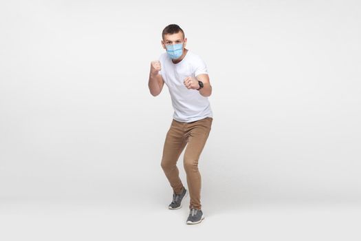 Full length portrait of aggressive young man in white shirt with surgical medical mask standing with boxing fists and ready to attack or defence. indoor studio shot, isolated on gray background.