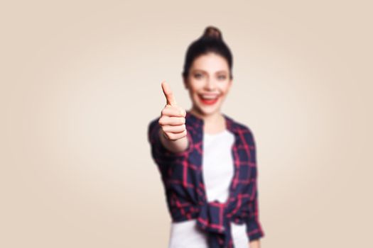 Young happy girl with casual style and bun hair thumbs up her finger, on beige blank wall with copy space looking at camera with toothy smile. focus on finger.