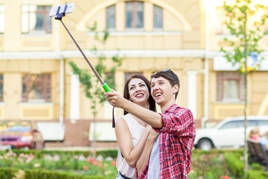 happy young tourists couple taking a selfie with smartphone on the monopod in city. The man is holding the stick and shooting looking at phone with happiness..