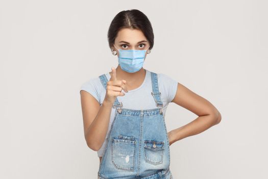 Portrait of serious young brunette woman with surgical medical mask in denim overalls standing looking at camera and warning or scolding. indoor studio shot isolated on gray background.