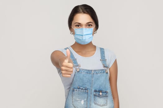 Its good, I like it. Portrait of satisfied young brunette woman with surgical medical mask in denim overalls standing, thumbs up and looking at camera . indoor studio shot isolated on gray background.
