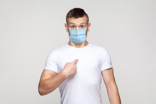 Who? me? Portrait of shocked young man in white shirt with surgical medical mask standing, poinitng himself and looking at camera with surprised face. indoor studio shot, isolated on gray background.