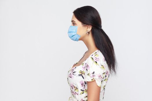 Profile side view portrait of calm young woman with medical mask in white dress standing . Protection against contagious disease, coronavirus. indoor studio shot isolated on gray background.