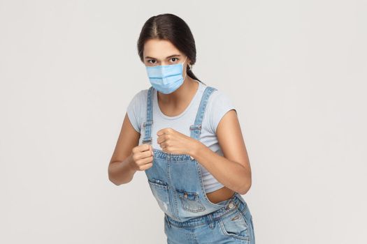 Portrait of angry mad young brunette woman with medical mask in denim overalls standing with boxing fists, looking and ready to attack or defence. indoor studio shot isolated on gray background.