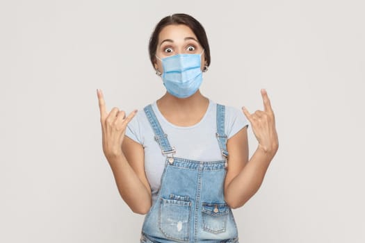 Yes. Portrait of amazed young brunette woman with medical mask in denim overalls standing with rock horns, looking at camera with surprised face. indoor studio shot isolated on gray background.