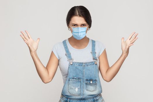 What? Portrait of shocked crazy young woman with surgical medical mask in denim overalls standing, raised arms and looking at camera with angry face. indoor studio shot isolated on gray background.