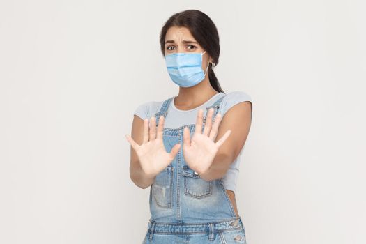 No, please wait, stop. Portrait of scared young brunette woman with medical mask in denim overalls standing and looking at camera with afraid face. indoor studio shot isolated on gray background.