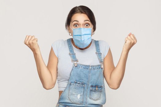 Wow, unbelievable news. Portrait of surprised young brunette woman with medical mask in denim overalls standing, celebrating and cheering her victory. indoor studio shot isolated on gray background.