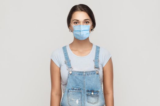 Portrait of young brunette woman with surgical medical mask in denim overalls standing and looking at camera. medical and health care concept. indoor studio shot isolated on gray background.