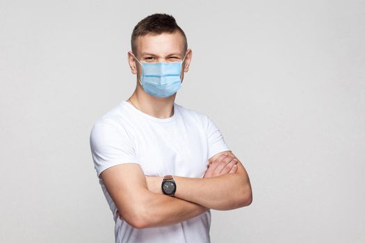 Portrait of funny young man in white shirt with surgical medical mask standing with crossed arms and looking at camera winking with funny face. indoor studio shot, isolated on gray background.