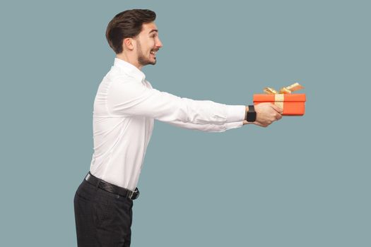 Profile side view of happy smiley bearded man in white shirt standing and holding red gift box, giving and side looking with toothy smile. Indoor, studio shot isolated on light blue background