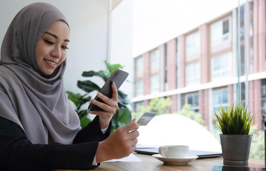 Beautiful young muslim woman with hijab sits at her office desk, holding a smartphone and credit card. Online payment, internet banking, online shopping concept. Close-up image.