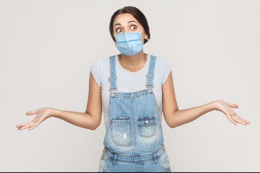 I don't know. Portrait of thoughtful young brunette woman with surgical medical mask in denim overalls standing raised arms and looking away. indoor studio shot isolated on gray background.