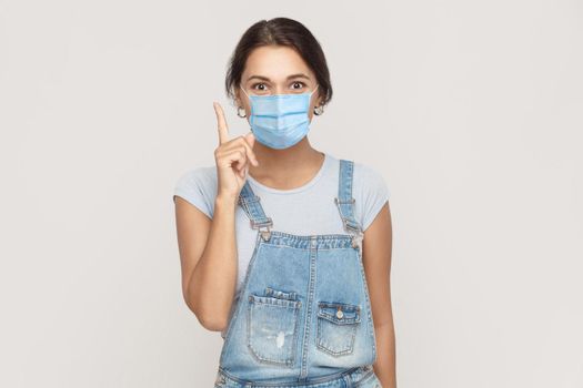 I have idea. Portrait of happy young brunette woman with surgical medical mask in denim overalls standing surprised and looking at camera smiling. indoor studio shot isolated on gray background.