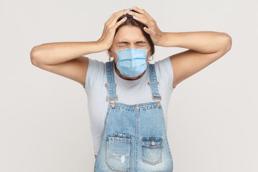 Problem, crisis, bad news or headache. Portrait of vexed young brunette woman with surgical medical mask in denim overalls standing and holding her head. indoor studio shot isolated on gray background