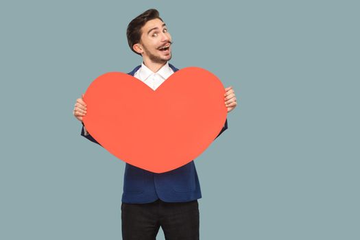 Handsome funny happy businessman in blue jacket and white shirt standing and holding red big heart shape and looking at camera with good feelings. Indoor, studio shot isolated on light blue background