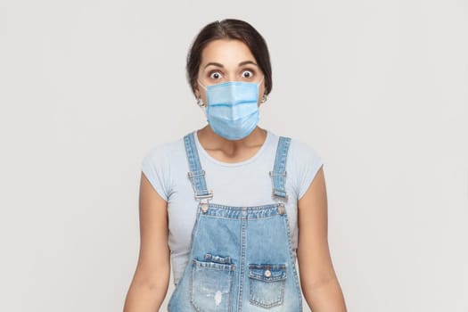 Portrait of shocked young brunette woman with surgical medical mask in denim overalls standing and looking at camera with big eyes and surprised. indoor studio shot isolated on gray background.