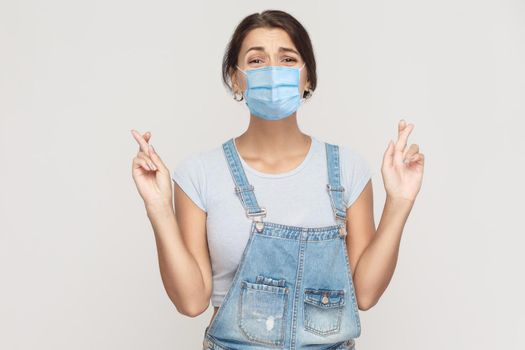 Portrait of young hopeful brunette woman with surgical medical mask in denim overalls standing crossed arms, looking at camera with wishful worry face. indoor studio shot isolated on gray background.