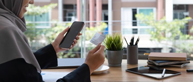 Beautiful young muslim woman with hijab sits at her office desk, holding a smartphone and credit card. Online payment, internet banking, online shopping concept. Close-up image.