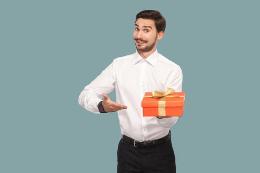 Funny happy bearded man in white shirt standing and holding red gift box, showing and looking at camera with toothy smile. Indoor, studio shot isolated on light blue background