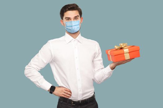 Young man with surgical medical mask in white shirt standing with hand on waist holding red gift box, looking at camera with satisfied face. health care concept. Indoor, isolated on blue background