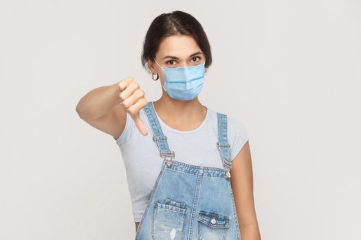 I don't like it. Portrait of dissatisfied young brunette woman with surgical medical mask in denim overalls standing thumbs down and looking at camera. indoor studio shot isolated on gray background.