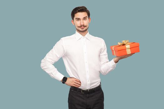 Happy bearded man in white shirt standing with hand on waist holding red gift box, looking at camera with satisfied face and smiling. Indoor, studio shot isolated on light blue background