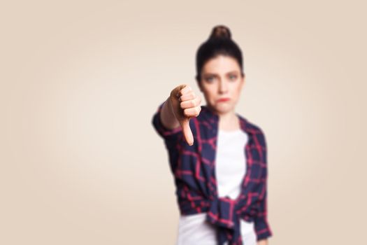 Dislike. Young unhappy upset girl with casual style and bun hair thumbs down her finger, on beige blank wall with copy space looking at camera with toothy smile. focus on finger.