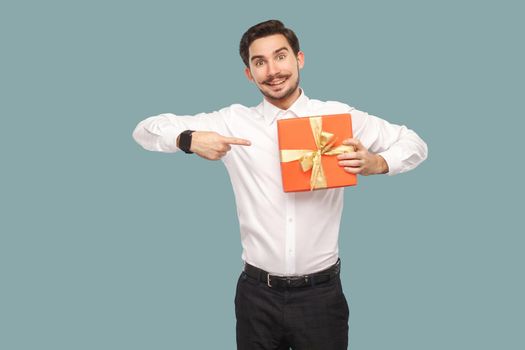Funny happy bearded man in white shirt standing and holding red gift box, pointing finger at present and looking at camera with toothy smile. Indoor, studio shot isolated on light blue background