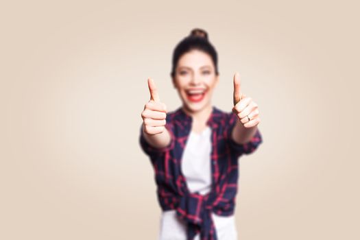 Young happy girl with casual style and bun hair thumbs up her finger, on beige blank wall with copy space looking at camera with toothy smile. focus on finger.