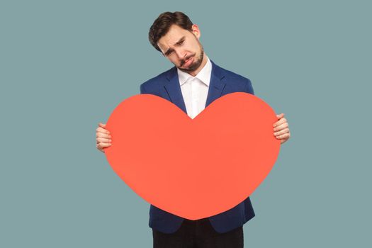 sad unhappy businessman in blue jacket and white shirt standing and holding red big heart shape and looking at camera with cry and broken heart. Indoor, studio shot isolated on light blue background.