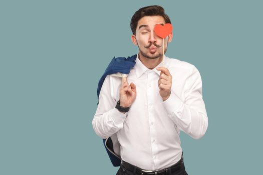 In love funny man in white shirt standing and holding red heart sticker in front of eye, kissing with closed eyes at camera. Indoor, studio shot isolated on light blue background.