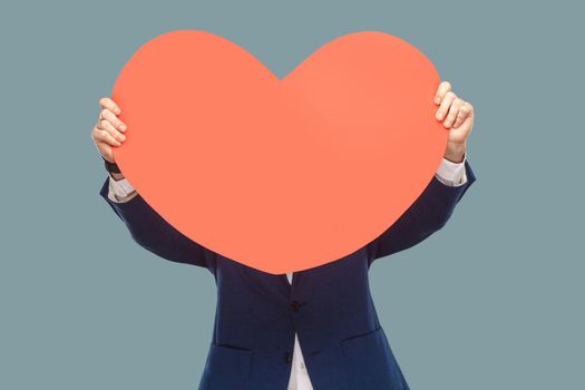 Handsome businessman in blue jacket standing and holding and covering red big heart shape in front of face for copy space. Indoor, studio shot isolated on light blue background.