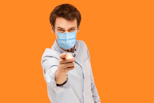 Hey you. Portrait of angry young worker man with surgical medical mask standing pointing and scolding at camera with mad face. indoor studio shot isolated on orange background.