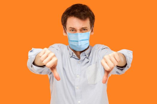 Portrait of sad young worker man with surgical medical mask standing thumbs down and looking at camera with dissatisfied face. indoor studio shot isolated on orange background.