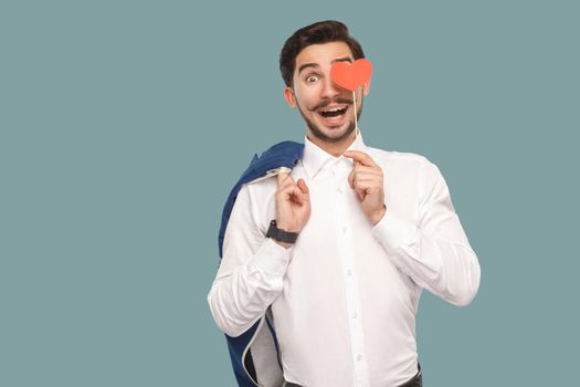 In love funny man in white shirt standing and holding red heart sticker and looking at camera with funny open mouth and amazed face and big eyes. Indoor, studio shot isolated on light blue background.