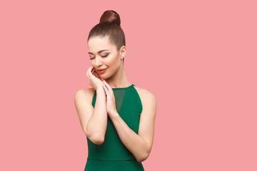 Beauty portrait of happy beautiful young woman with bun hairstyle and makeup in green dress standing with closed eyes, touching her face and smiling. indoor studio shot, isolated on pink background.